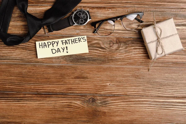 Top view of gift box, greeting card with lettering happy fathers day and mens black tie, wristwatch and glasses on wooden background — Stock Photo