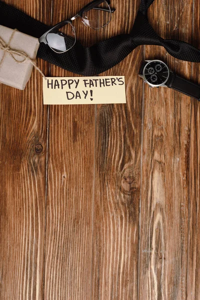 Top view of gift box, greeting card with lettering happy fathers day and mens black tie, wristwatch, glasses and copy space on wooden background — Stock Photo