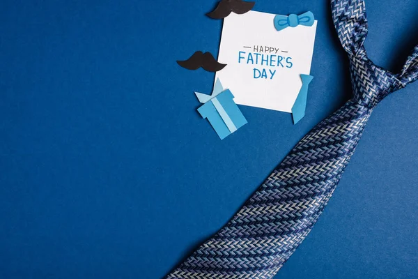 Vista superior de la tarjeta de felicitación con letras feliz día de los padres, elementos de decoración artesanal de papel y mens corbata sobre fondo azul - foto de stock