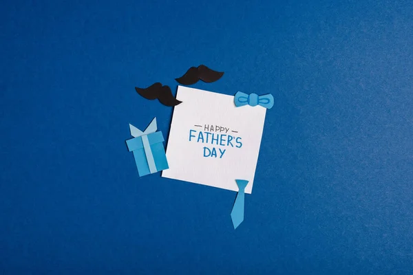 Vista superior de la tarjeta de felicitación con letras feliz día de los padres y elementos de decoración artesanal de papel sobre fondo azul - foto de stock