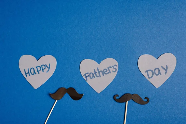 Vista superior de cartón decorativo bigote falso y papel cortado corazones grises con letras feliz día de los padres sobre fondo azul - foto de stock