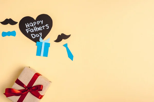 Vista superior de la caja de regalo, elementos decorativos de papel y tarjeta en forma de corazón de saludo con letras feliz día de los padres sobre fondo beige - foto de stock