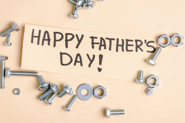 Vista superior de tuercas metálicas y pernos y tarjeta de felicitación beige con letras escritas a mano feliz día de los padres sobre fondo beige - foto de stock