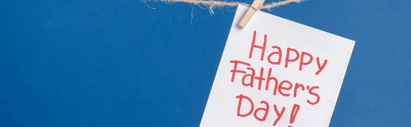 Panoramic shot of white paper card with red lettering happy fathers day hanging on rope with clothespins isolated on blue — Stock Photo