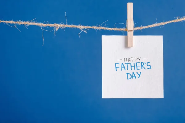 Tarjeta de felicitación blanca con letras feliz día de los padres colgando de la cuerda con pinzas de ropa aisladas en azul - foto de stock