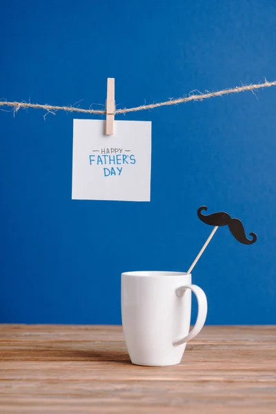 Selektiver Fokus der weißen Tasse mit Zierpapier falscher Schnurrbart und weiße Grußkarte mit Schriftzug Happy Vatertag isoliert auf blau — Stockfoto