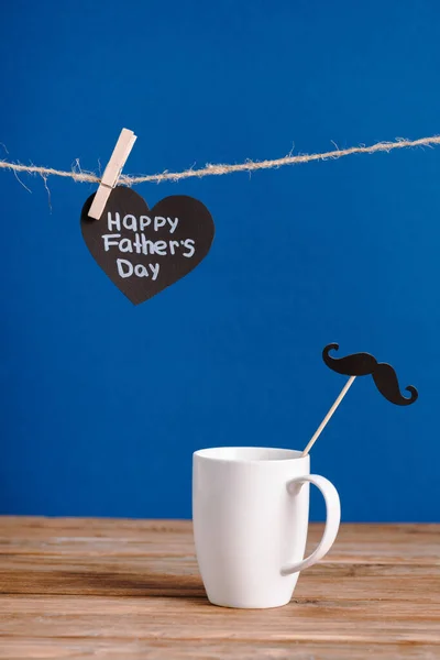 Selektiver Fokus der weißen Tasse mit Zierpapier falscher Schnurrbart und schwarzer Grußkarte mit Schriftzug Happy Vatertag isoliert auf blau — Stockfoto