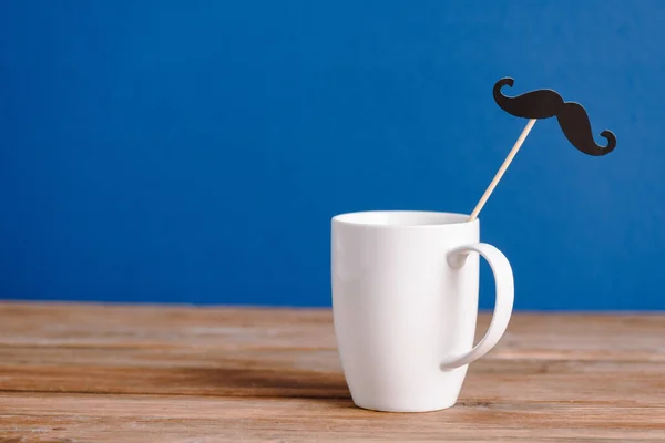 Taza blanca y papel decorativo negro bigote falso en la superficie de madera aislado en azul - foto de stock