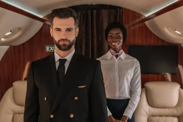 Избирательный фокус уверенного пилота и улыбающейся африканской стюардессы, смотрящей в камеру в частном самолете — стоковое фото