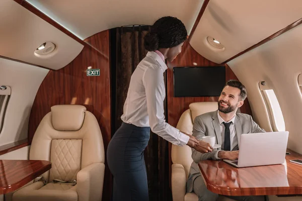 Junge afrikanisch-amerikanische Stewardess gibt lächelnden Geschäftsmann, der im Flugzeug am Laptop sitzt, eine Tasse Kaffee — Stockfoto