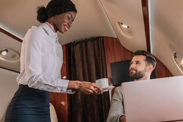 Lächelnde afrikanisch-amerikanische Stewardess, die einem hübschen Geschäftsmann, der im Privatjet am Laptop arbeitet, eine Tasse Kaffee reicht — Stockfoto