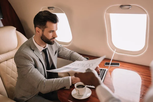 Abgeschnittene Ansicht einer afrikanisch-amerikanischen Stewardess, die einem Geschäftsmann, der im Privatjet am Laptop arbeitet, Zeitung schenkt — Stockfoto