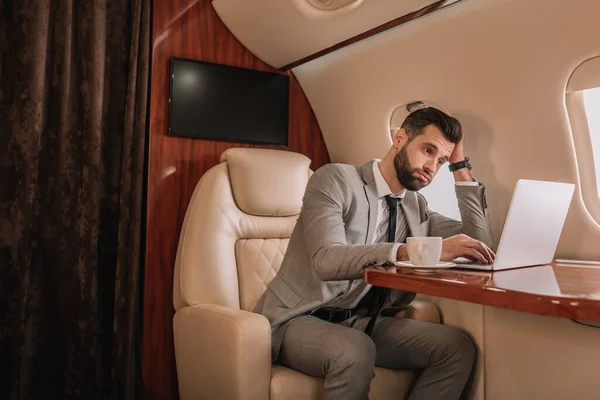 Bored businessman touching head while working on laptop in private plane — Stock Photo