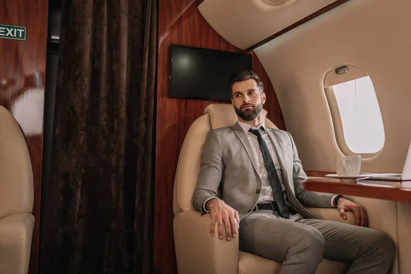 Guapo hombre de negocios viajando en avión privado mientras está sentado cerca de la mesa con una taza de café - foto de stock