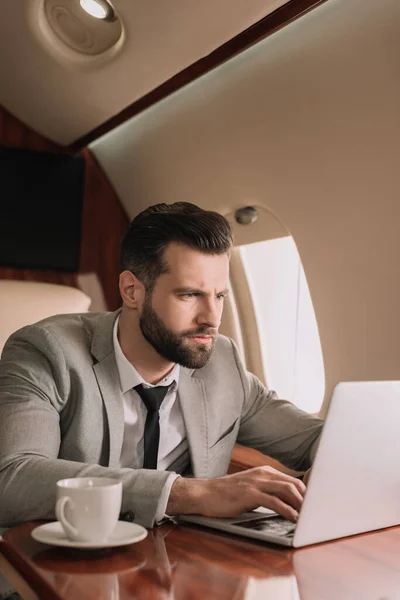 Hombre de negocios concentrado escribiendo en el ordenador portátil cerca de la taza de café en avión privado - foto de stock