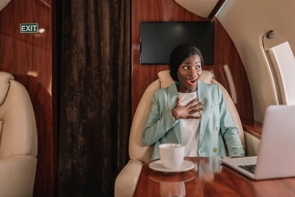 Шокована афроамериканська бізнес-леді в приватному літаку торкаючись грудей, дивлячись в отвір — стокове фото