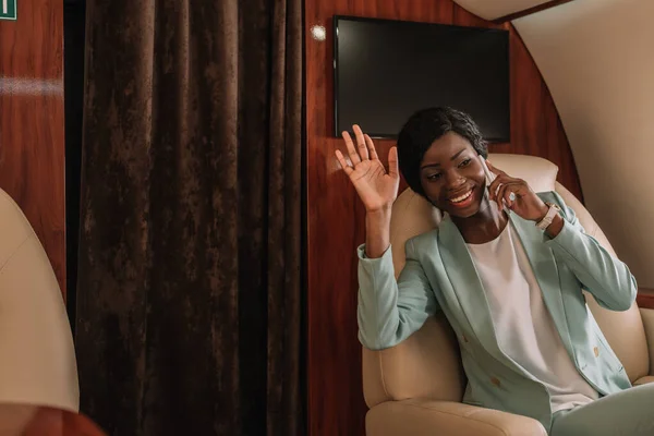 Счастливая африканская американская деловая женщина машет рукой, разговаривая по смартфону в частном самолете — стоковое фото