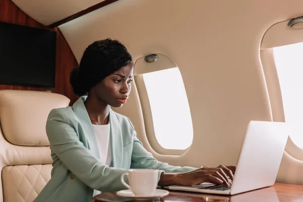 Atractiva, confiada mujer de negocios afroamericana que trabaja en el ordenador portátil en jet privado - foto de stock