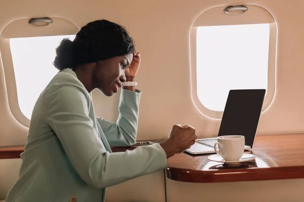 Улыбающаяся африканская американская деловая женщина рядом с ноутбуком с чистым экраном и чашкой кофе в частном самолете — стоковое фото