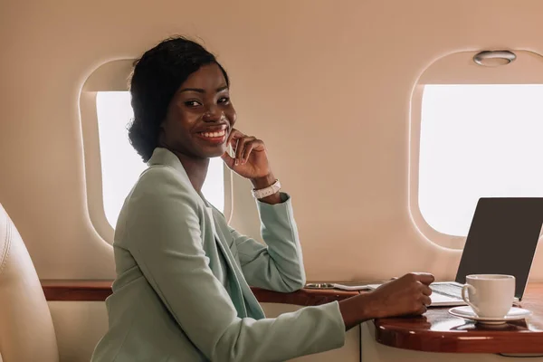 Attractive african american businesswoman smiling at camera near laptop and coffee cup in private jet — Stock Photo