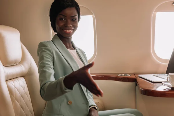 Glückliche afrikanisch-amerikanische Geschäftsfrau zeigt Grußgeste mit ausgestreckter Hand im Privatjet — Stockfoto