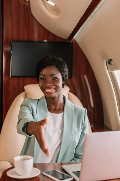 Sonriente mujer de negocios afroamericana mostrando gesto de saludo con la mano extendida en jet privado - foto de stock