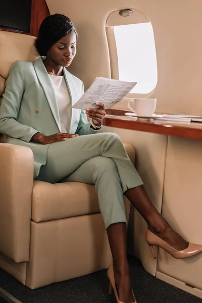 Atractiva mujer de negocios afroamericana leyendo documento en avión privado - foto de stock