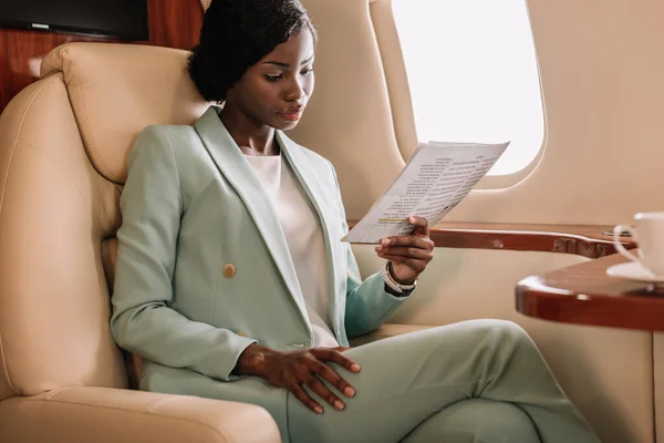 Mujer de negocios afroamericana concentrada leyendo documento en jet privado - foto de stock