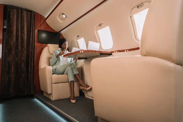 Confident african american businesswoman drinking coffee and reading newspaper in private jet — Stock Photo