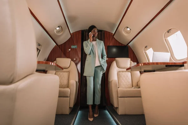 Enfoque selectivo de atractiva mujer de negocios afroamericana hablando en el teléfono inteligente y cogido de la mano en el bolsillo - foto de stock