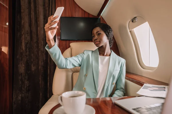 Atractiva mujer de negocios afroamericana tomando selfie en smartphone en avión privado - foto de stock