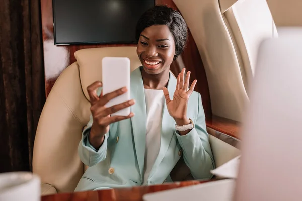 Enfoque selectivo de la feliz mujer de negocios afroamericana agitando la mano durante la videollamada en el teléfono inteligente - foto de stock