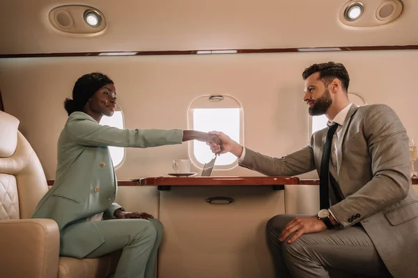 Vista laterale di attraente donna d'affari africana americana che stringe la mano con l'uomo d'affari in aereo — Foto stock
