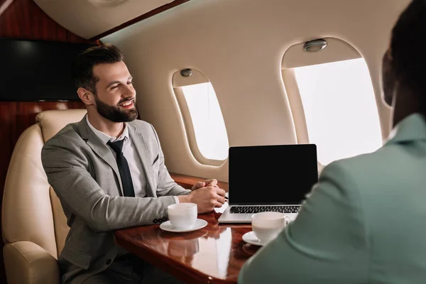 Enfoque selectivo de hombre de negocios sonriente mirando a la mujer de negocios afroamericana cerca de la computadora portátil con pantalla en blanco - foto de stock