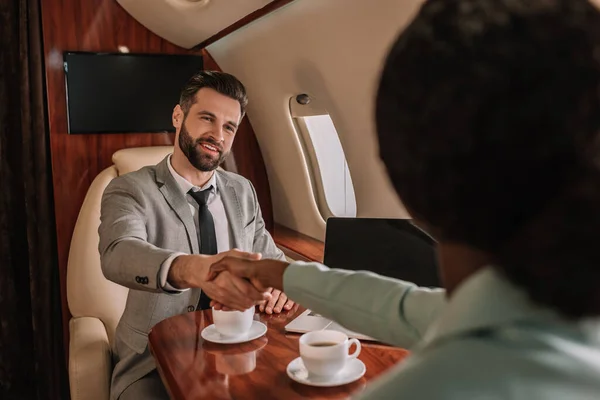 Selektiver Fokus eines lächelnden Geschäftsmannes beim Händeschütteln mit einer afrikanisch-amerikanischen Geschäftsfrau im Flugzeug — Stockfoto