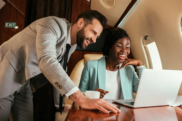 Sonriente hombre de negocios de pie cerca de la feliz mujer de negocios afroamericana mientras que tiene videollamada en jet privado - foto de stock
