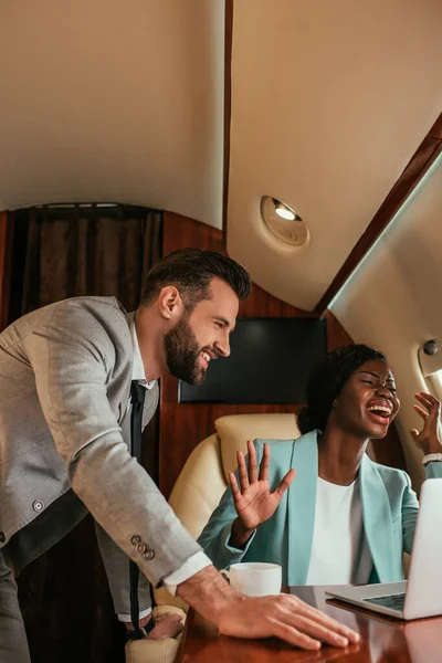 Homme d'affaires souriant debout près excité femme d'affaires afro-américaine agitant la main tout en ayant appel vidéo en jet privé — Photo de stock