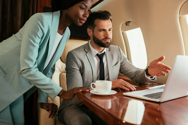 Serious businessman pointing with finger at laptop near african american businesswoman — Stock Photo