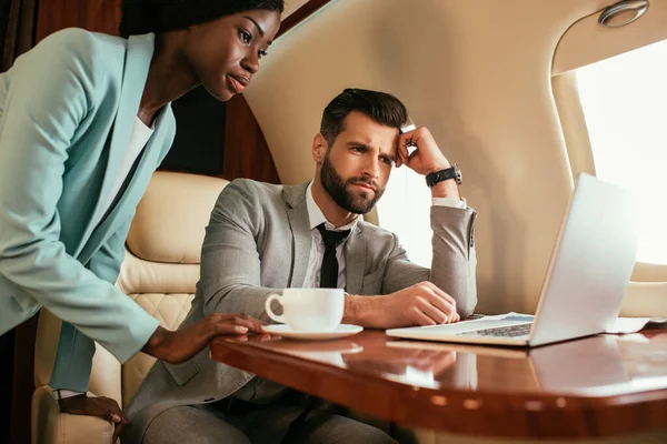 Homme d'affaires sérieux et femme d'affaires afro-américaine à la recherche d'un ordinateur portable en jet privé — Photo de stock