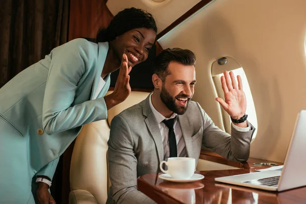 Souriant hommes d'affaires muiticulturels agitant les mains tout en ayant chat vidéo sur ordinateur portable en jet privé — Photo de stock