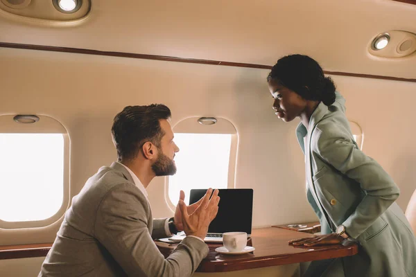 Confiado hombre de negocios afroamericano de pie cerca de hombre de negocios sentado a la mesa en jet privado - foto de stock