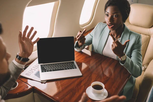 Mujer de negocios afroamericana enojada mostrando los dedos medios mientras está sentado frente a hombre de negocios en jet privado - foto de stock