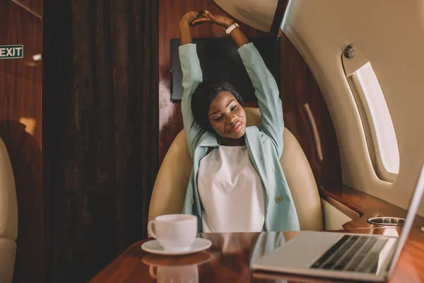 Sorridente mulher de negócios afro-americana que se estende com as mãos levantadas perto de laptop e xícara de café em jato privado — Fotografia de Stock