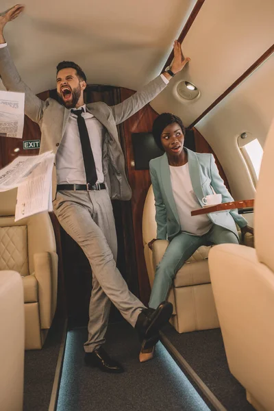 Emocionado hombre de negocios gritando y tocando techo mientras asustada mujer de negocios afroamericana sentado en jet privado - foto de stock