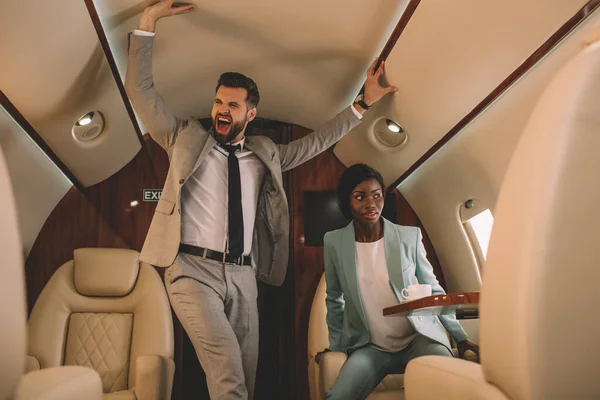 Excited businessman screaming and touching ceiling while scared african american businesswoman sitting in private jet — Stock Photo