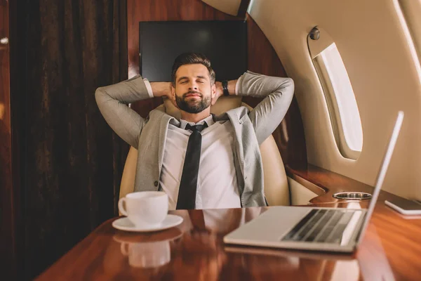 Молодий бізнесмен відпочиває руками за голову і закриті очі біля ноутбука і чашки кави в приватному літаку — стокове фото