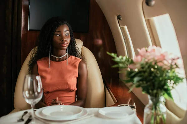 Enfoque selectivo de la mujer afroamericana soñadora sentada en la mesa servida en avión privado - foto de stock