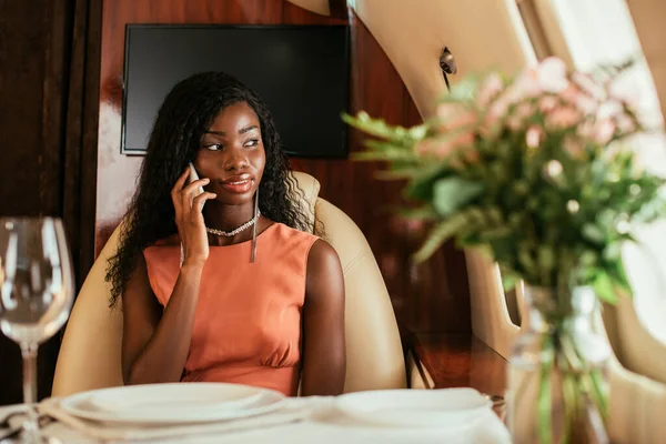 Enfoque selectivo de la atractiva mujer afroamericana hablando en el teléfono inteligente en avión privado - foto de stock