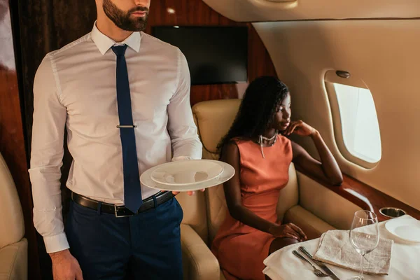 Vista recortada del administrador de aire sosteniendo placas cerca de la mujer afroamericana pensativa sentada a la mesa servida en avión - foto de stock