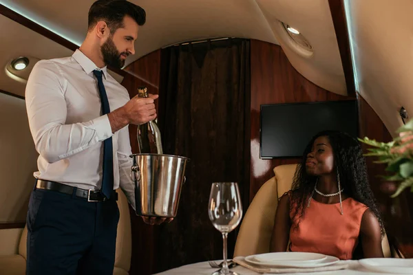 Schöner Flugbegleiter mit Champagner-Eimer und Flasche in der Nähe attraktiver Afroamerikanerin im Privatjet — Stockfoto